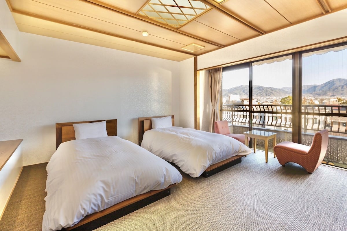 A hotel room with two beds, a small table, a chair, and a balcony with a view of mountains.