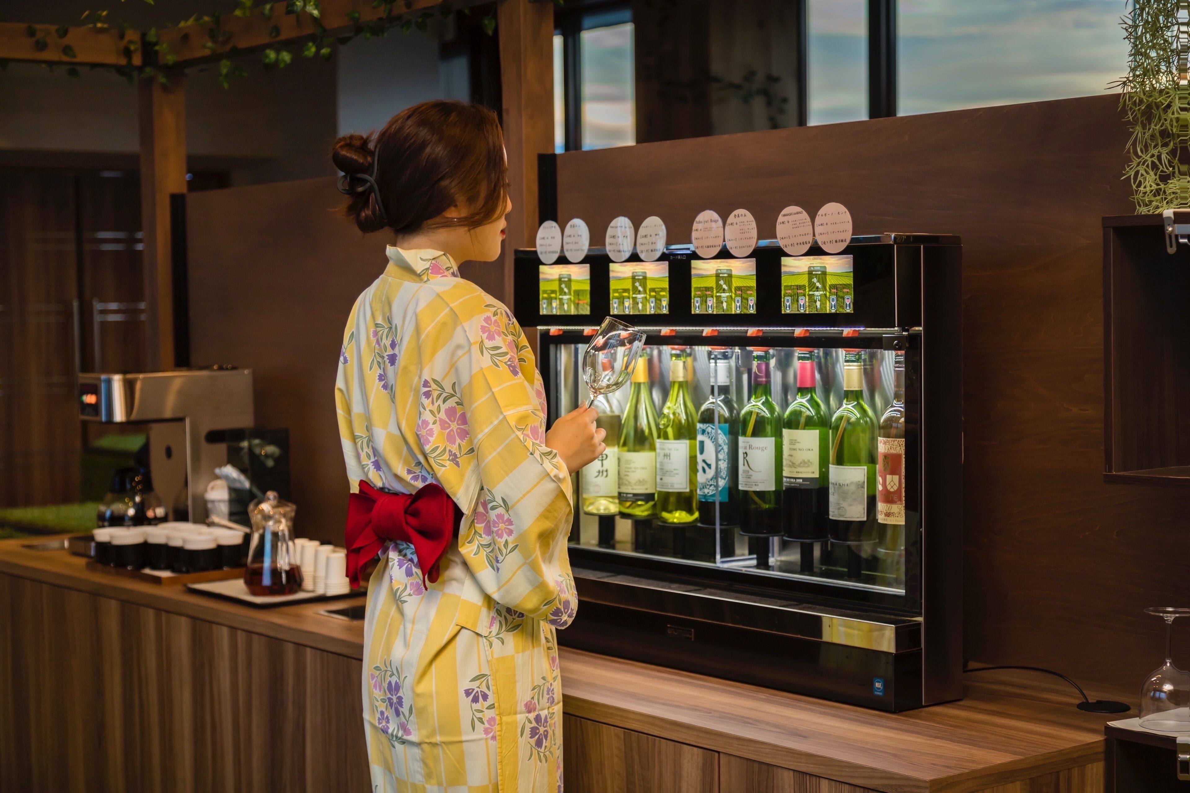 Une personne vêtue d’une robe à motifs floraux se tient devant un comptoir où se trouve un réfrigérateur à vin, dans lequel sont exposées différentes bouteilles.