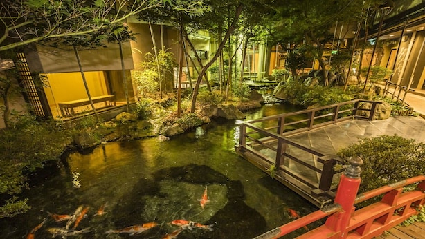 Ein Teich mit Koi-Karpfen, mit einer Holzbrücke, umgeben von üppigem Grün und traditioneller Architektur.
