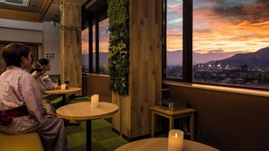 Ein Zimmer mit Holzwänden, einem großen Fenster mit Blick auf einen Sonnenuntergang und Berge, und einem kleinen Tisch mit einer Kerze.