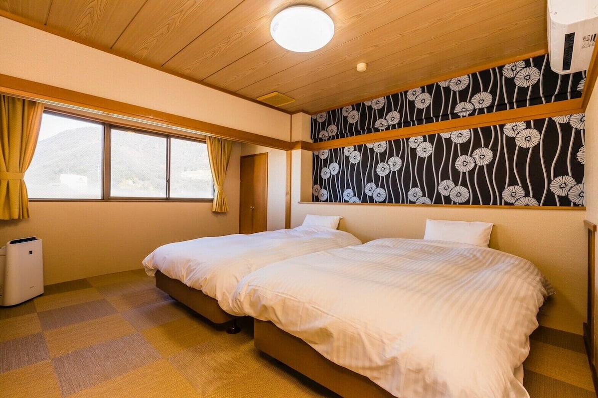 A hotel room with two beds, a large window with curtains, a ceiling fan, and a wall with a floral pattern.