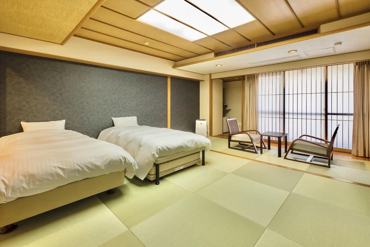 A hotel room with two beds, a chair, a table, and a large window with curtains.