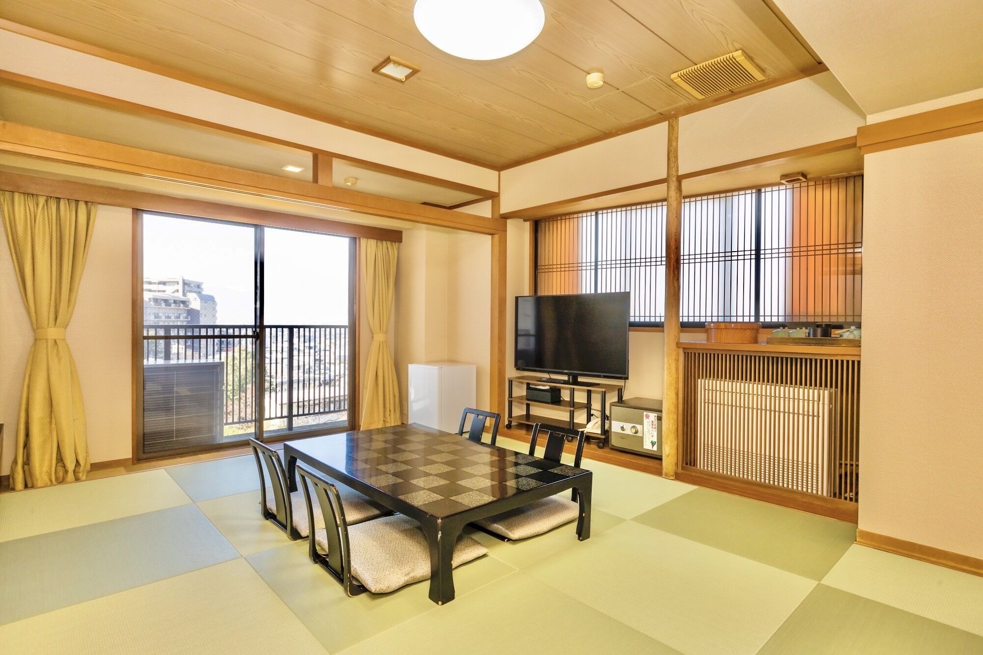 A room with tatami mat flooring, a low table with cushions, a television, and a sliding door to a balcony.