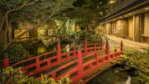 Ein traditioneller japanischer Garten mit einer roten Brücke über einem Teich, umgeben von üppiger Vegetation und traditioneller Architektur.