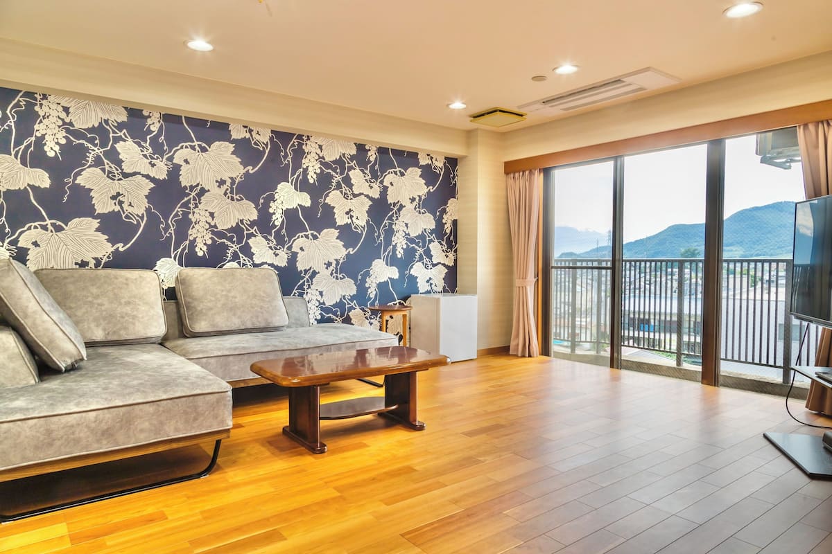 A modern living room with a sofa, a wooden coffee table, and a large window with a view of a balcony and mountains.