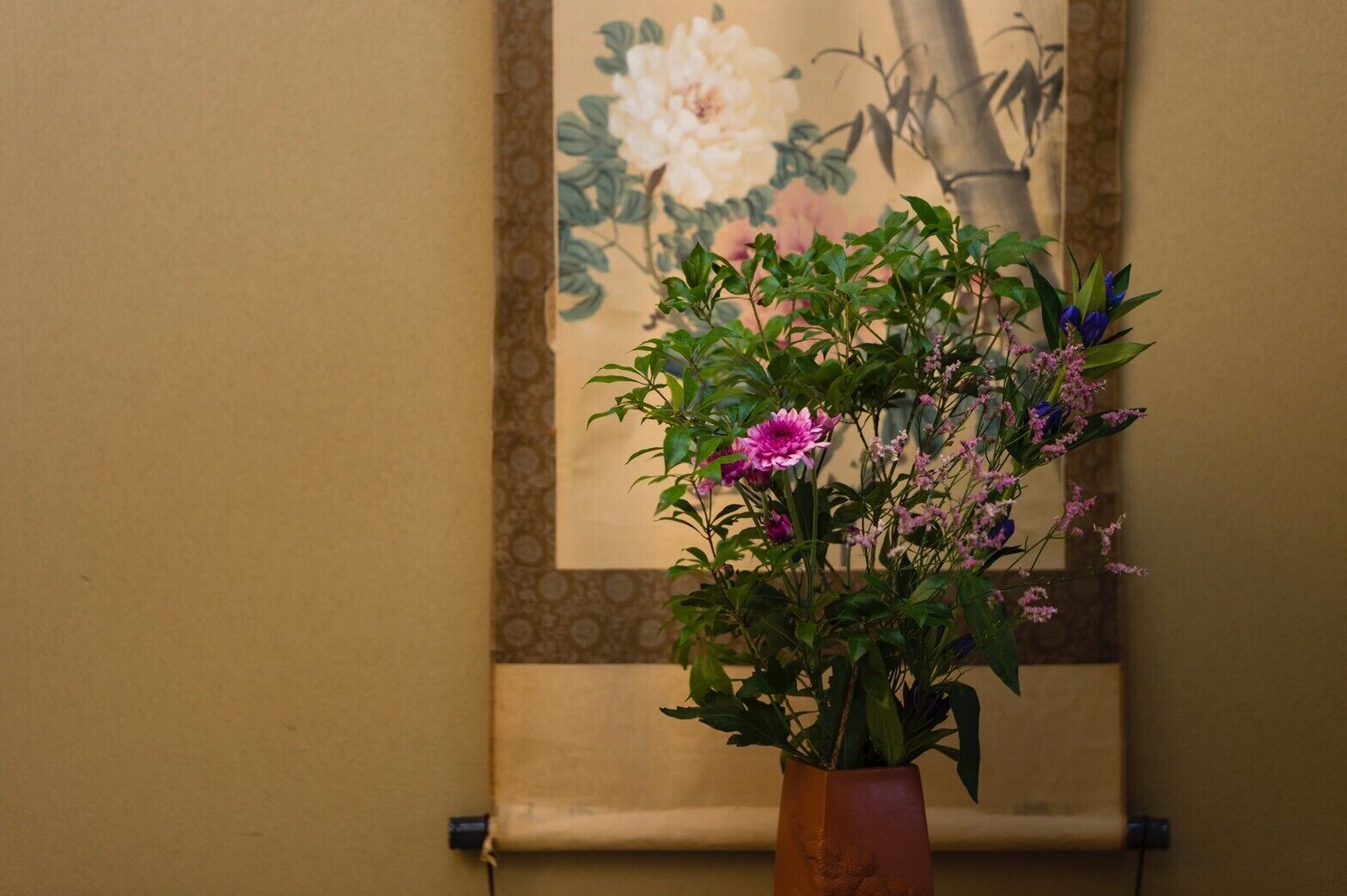 A potted plant with flowers in a room with a patterned wall and a painting.