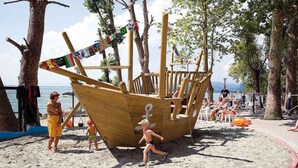 Eine hölzerne, schiffsförmige Spielanlage am Strand, mit Bäumen und einem Zaun im Hintergrund.