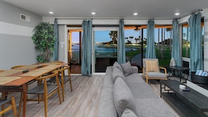 A modern living room with a large gray sofa, wooden dining table with woven placemats, and a view of a golf course through large windows with blue curtains. - Bayfront | Ultimate Family Fun | AC | Parking (San Diego)
