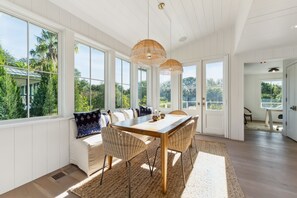 A dining area with a wooden table and wicker chairs, large windows with a view of greenery, and a ceiling with exposed beams. - Incredible Sullivan's Island Home with Pool - Monthly Rental Only (Sullivans Island)