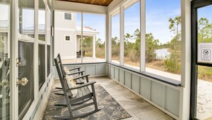 A covered porch with a rocking chair, large windows, and a view of a house and landscape. - 3BR Home @ Summer Salt • Sleeps 13 • Porches, Pool & Near Beach (Orange Beach)