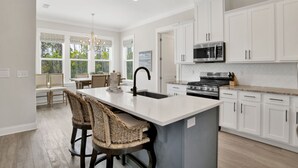 A modern kitchen with white cabinetry, a large island with bar stools, and a view of the outdoors through large windows. - 3BR Home @ Summer Salt • Sleeps 13 • Porches, Pool & Near Beach (Orange Beach)
