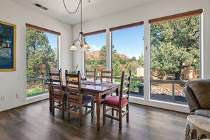 Uma área de refeições com mesa e cadeiras de madeira, grandes janelas com vista para árvores e montanhas, e um lustre.