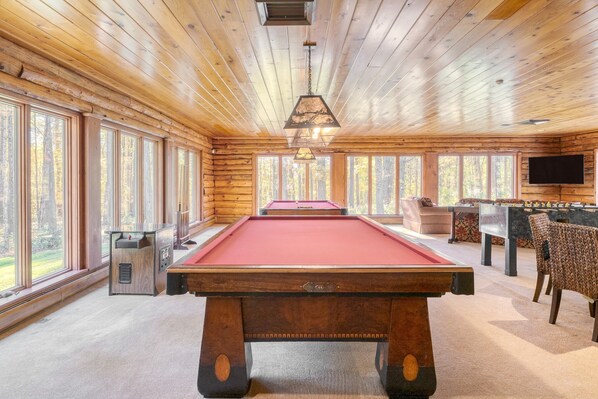 A pool table in a room with wooden walls and large windows, a television, and a seating area.
