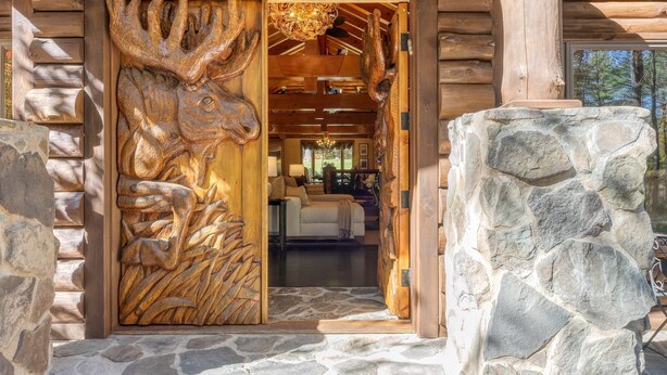 A wooden cabin entrance with a carved moose head, stone walls, and a cozy interior with a sofa and chandelier. - Moose Lodge by Avantstay Luxe Berkshires Escape (Great Barrington)