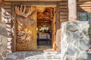 A wooden cabin entrance with a carved moose head, stone walls, and a cozy interior with a sofa and chandelier.