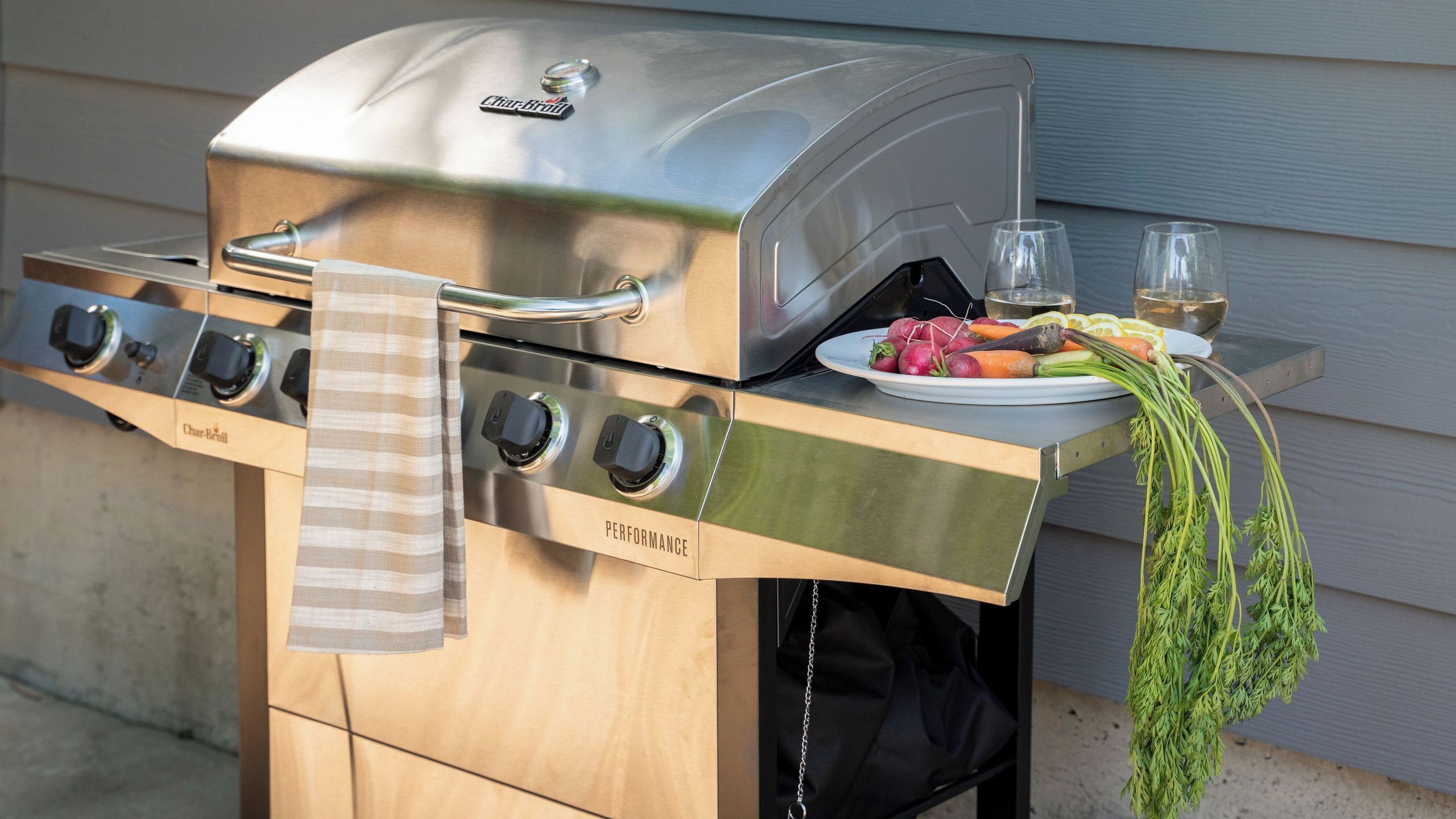 A Char-Broil Performance grill with a stainless steel finish, a towel hanging on the handle, and a plate of vegetables and lemons on the side.