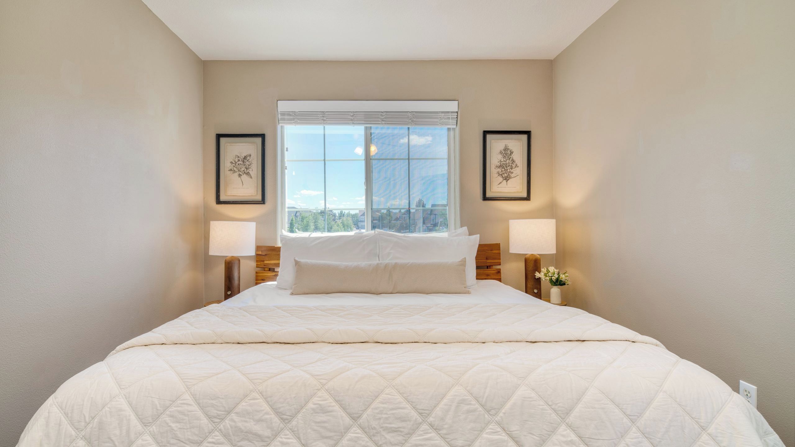 Een hotelkamer met een groot bed, twee bedlampjes, een raam met uitzicht op de stad en twee foto's in lijsten aan de muur.