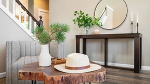 Quarto com sofá cinzento, mesa de madeira com chapéu, vaso branco com folhagem, espelho e console com velas.