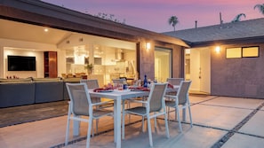 A modern outdoor dining area with a table and chairs, a kitchen in the background, and a sunset sky.