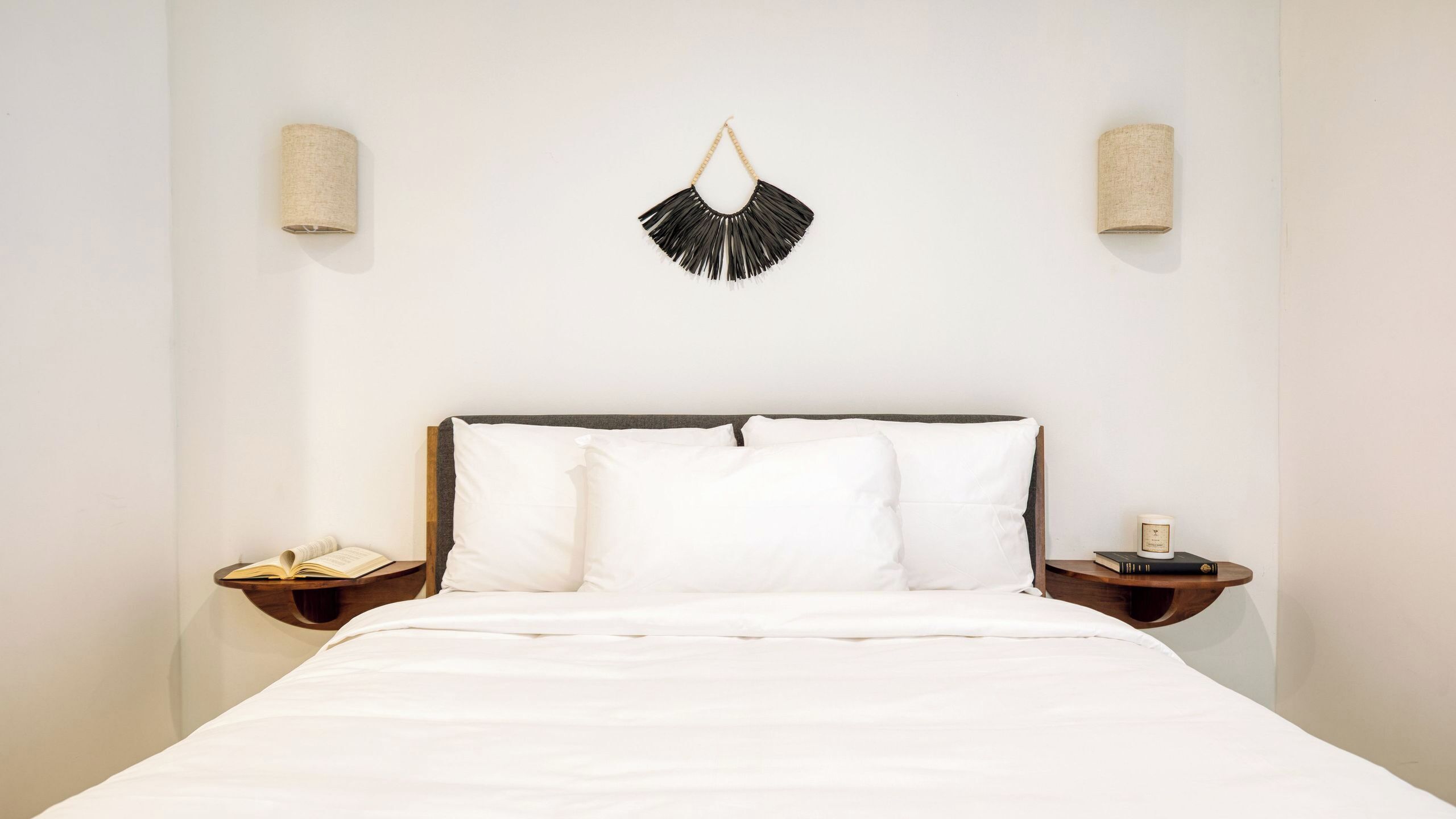 A bed with white bedding, a wooden headboard, two bedside tables with books and a candle, and wall-mounted sconces.