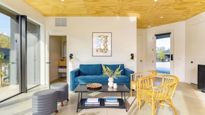 A modern living room with a blue sofa, a coffee table, a wicker chair, and a large window. - Blackhaus C & D Buyout by Avantstay Sleeps 16 (Palm Springs)
