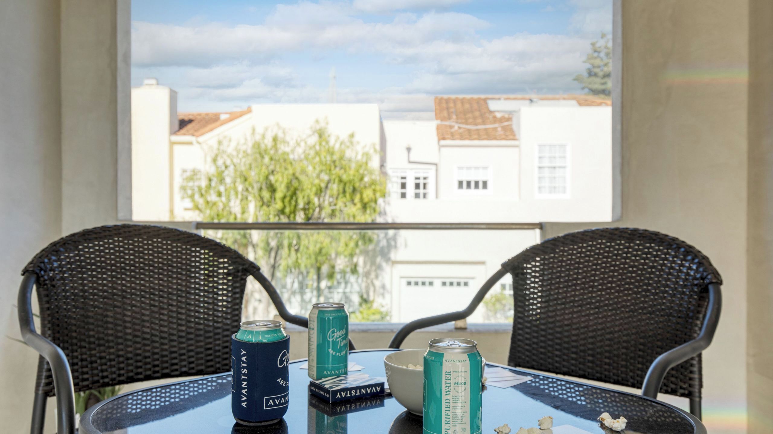 A balcony with two wicker chairs, a table with a glass top, and two cans of beverage.