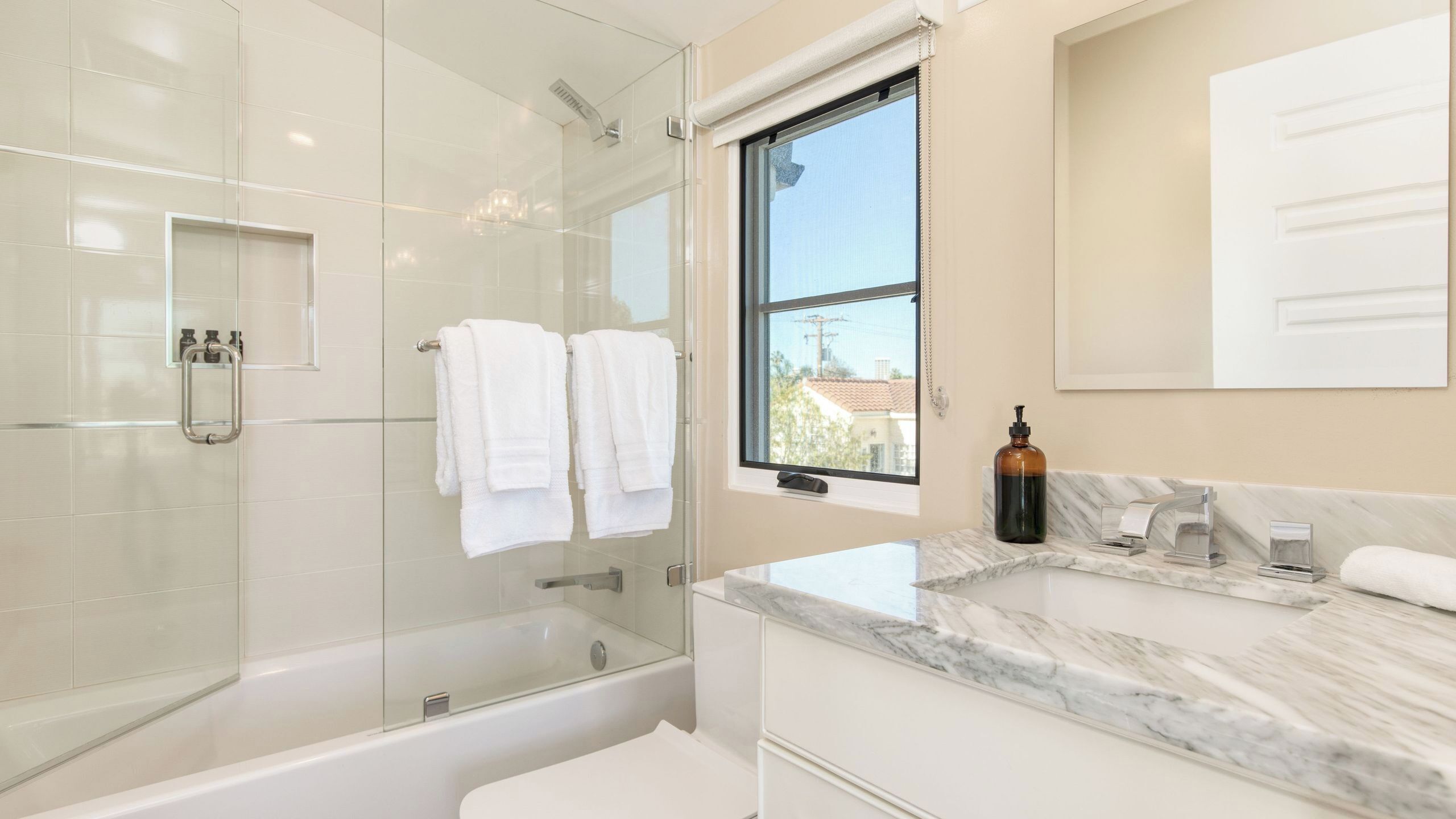 A modern bathroom with a glass shower enclosure, a bathtub, a marble vanity, and a window providing a view.