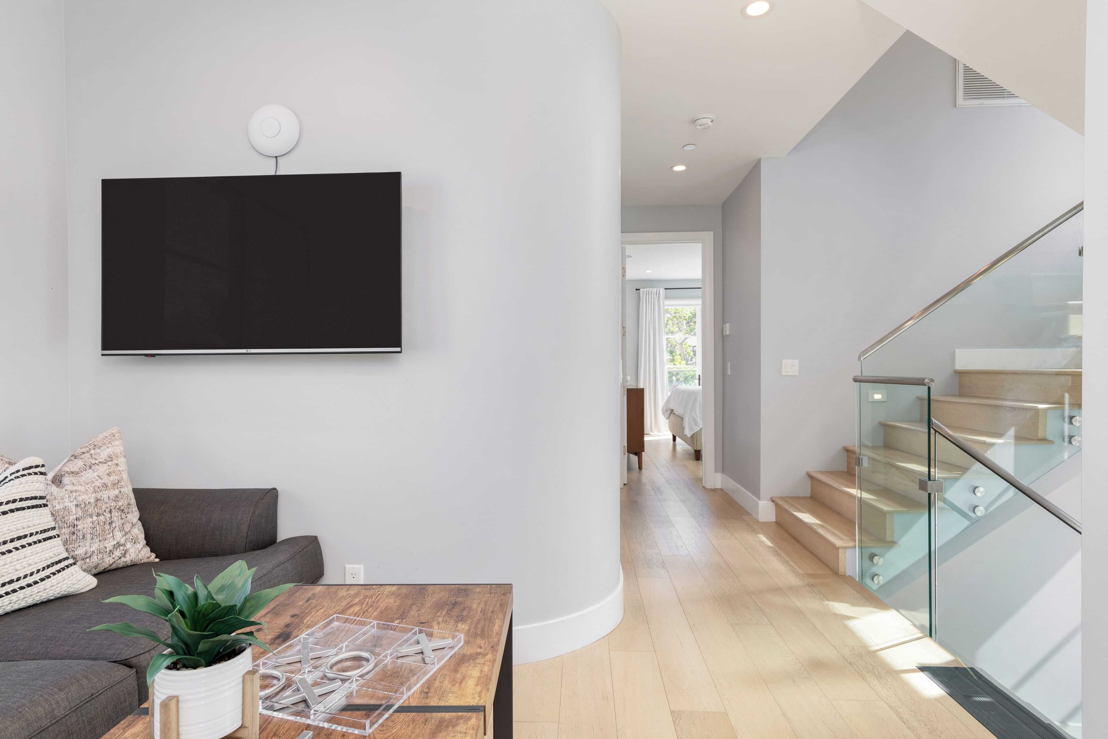 A modern living room with a flat-screen TV, a sofa, a coffee table with a plant, and a staircase with glass railings.