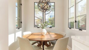 Uma sala de jantar com uma mesa de madeira redonda, seis cadeiras, um vaso branco com folhagem e um lustre único.