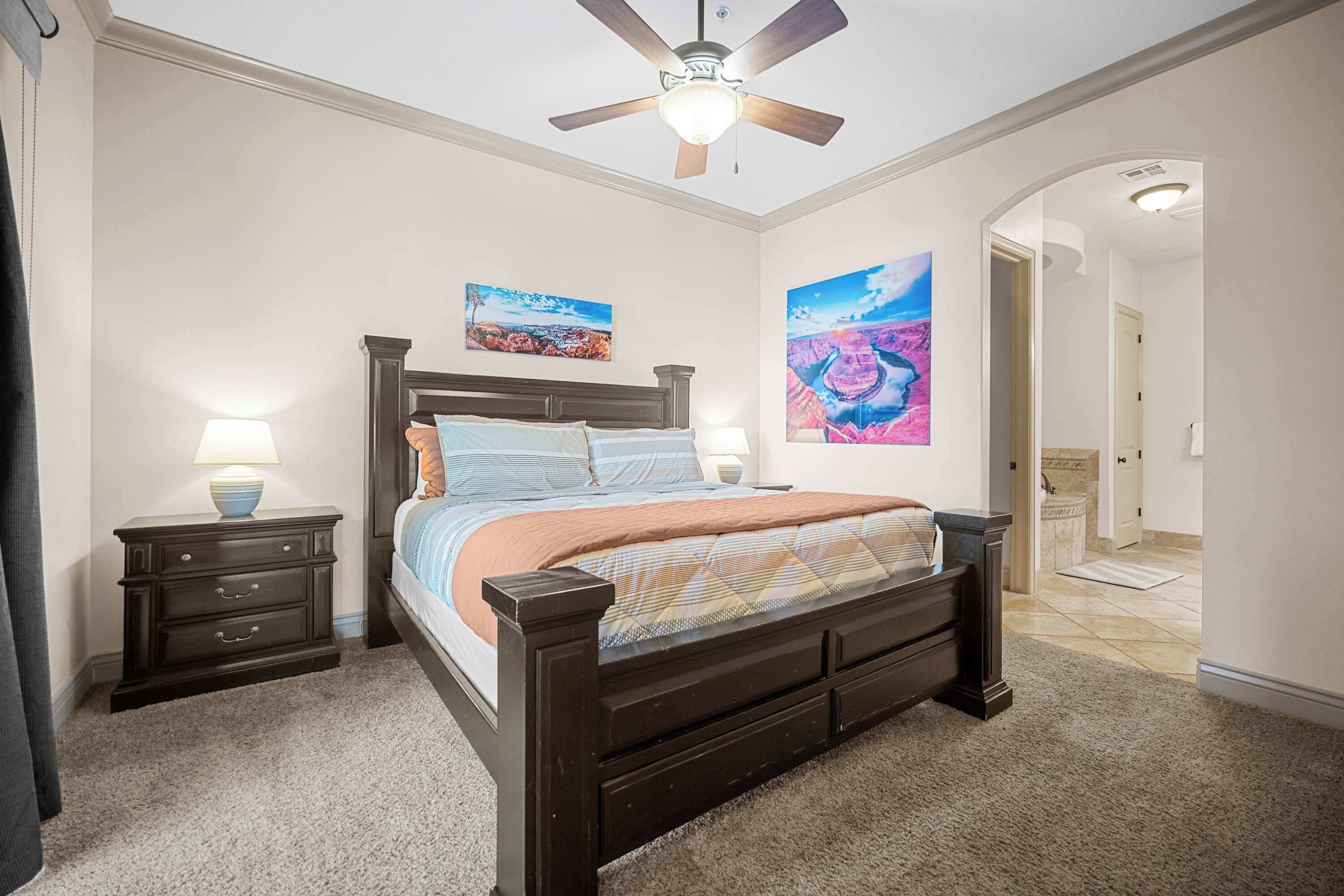 Quarto com uma cama de madeira escura, duas mesas de cabeceira com luminárias, um ventilador de teto e duas grandes pinturas de paisagens.