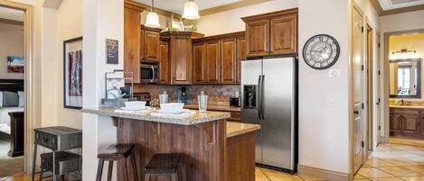 A kitchen with wooden cabinets, a stainless steel refrigerator, a large island with stools, pendant lighting, and a clock on the wall.
