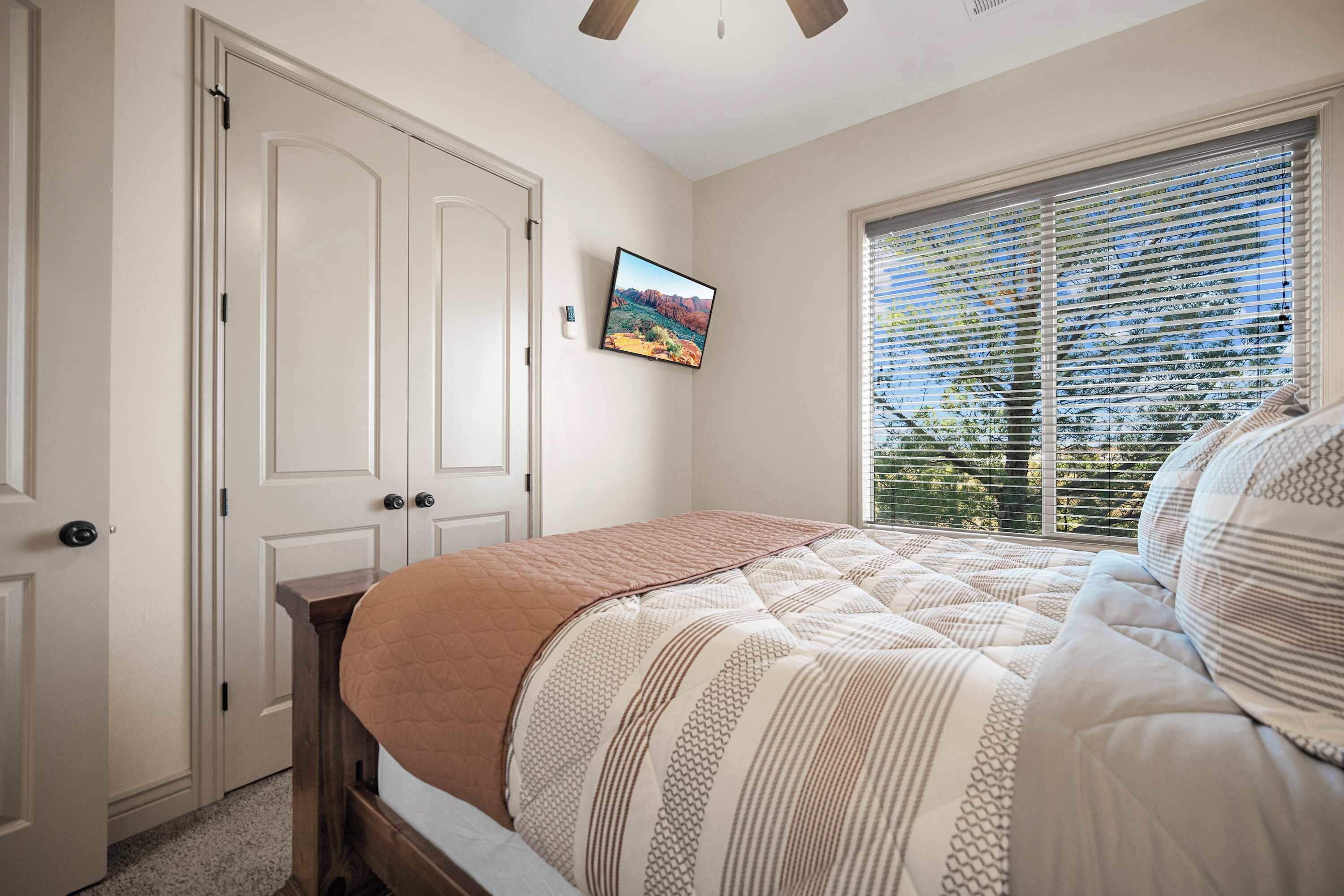 A bedroom with a bed, a window with blinds, a door, and a wall-mounted TV.