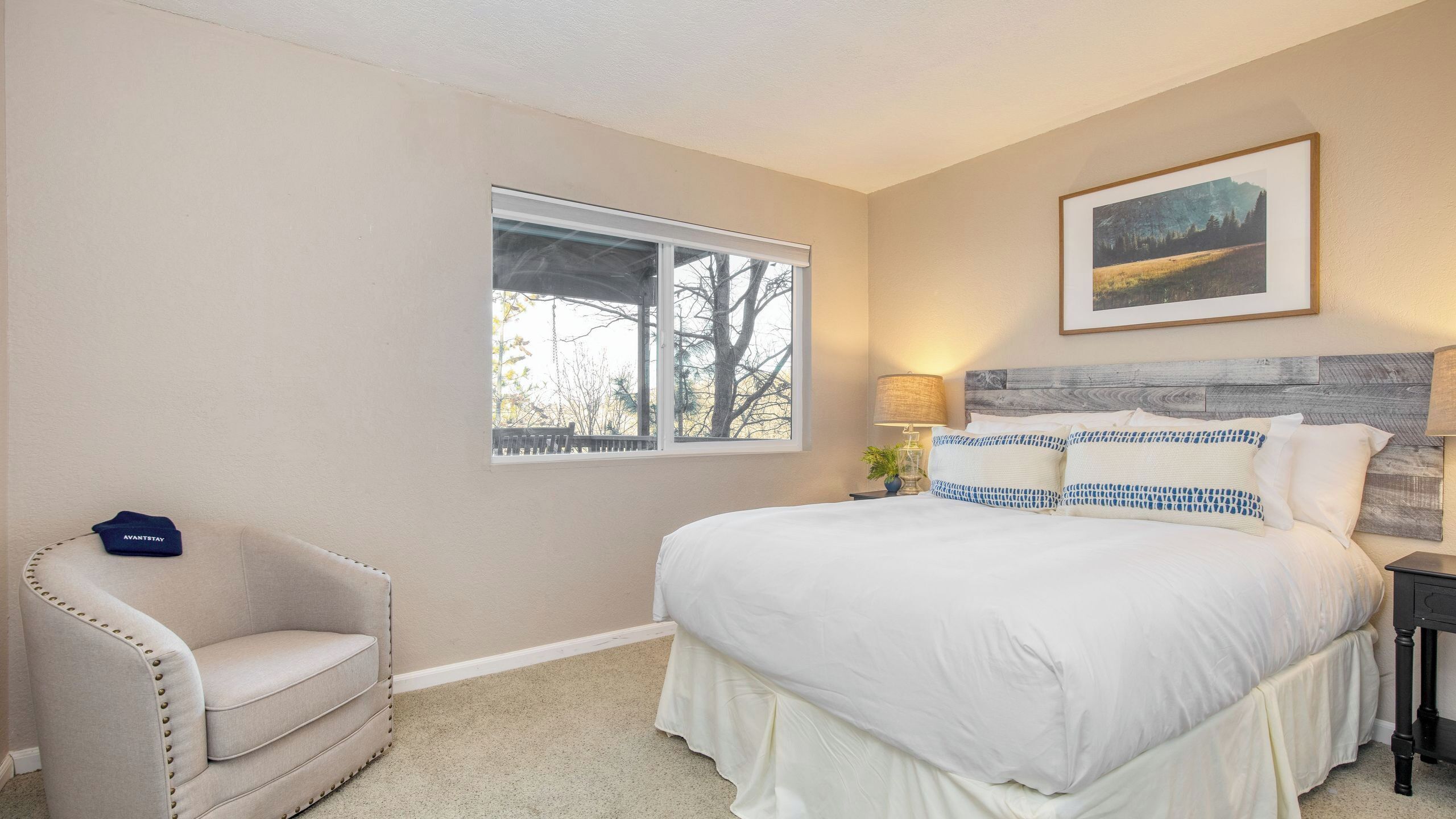 A bedroom with a bed, a chair, a window, a lamp, and a painting on the wall.