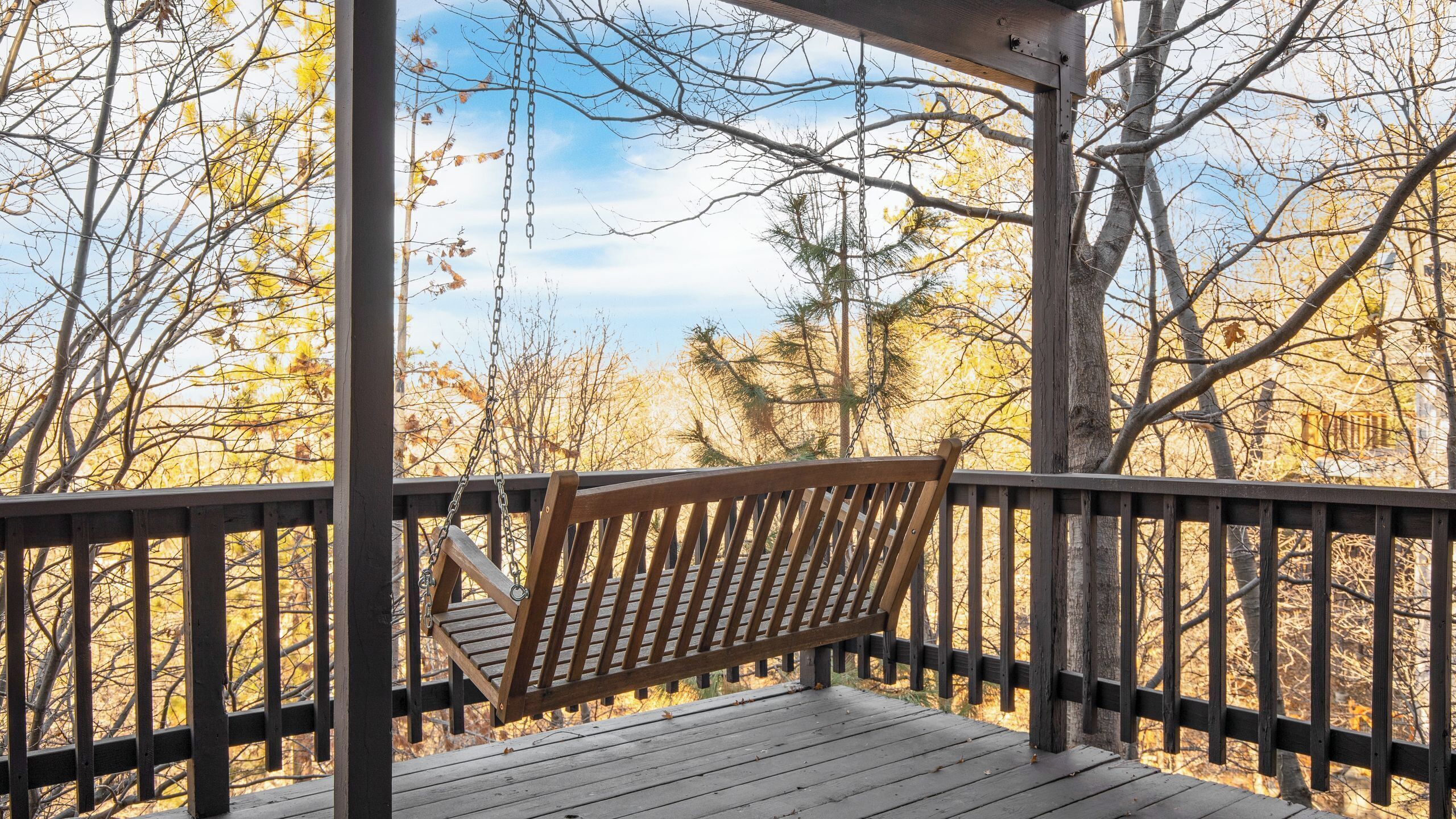 A wooden deck with a swing, surrounded by trees with autumn foliage.