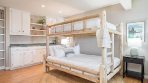 Un lit superposé avec une literie blanche, une petite table de chevet avec une lampe, un tableau encadré au mur et un coin cuisine avec des meubles blancs et un plan de travail.