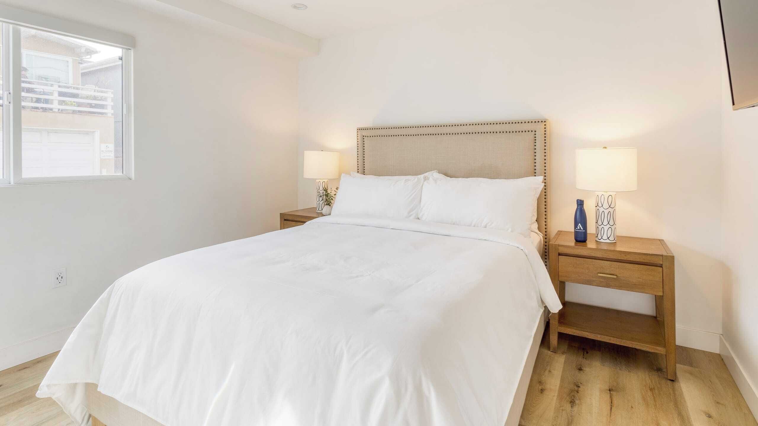 A neatly made bed with white linens, two bedside tables with lamps, and a window with a view of a building outside.