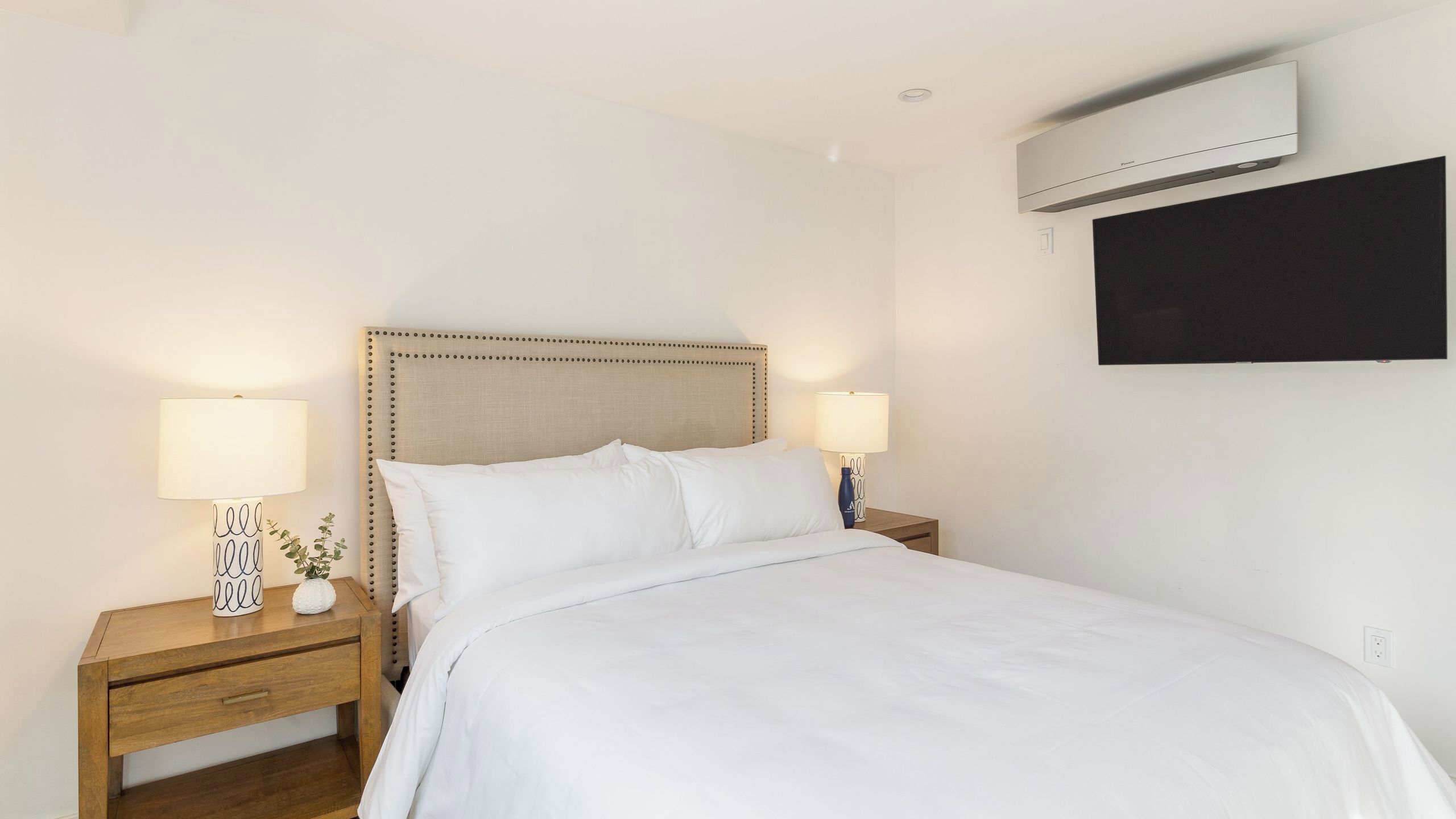 A hotel room with a bed, two bedside tables with lamps, a wall-mounted air conditioner, and a blackboard.