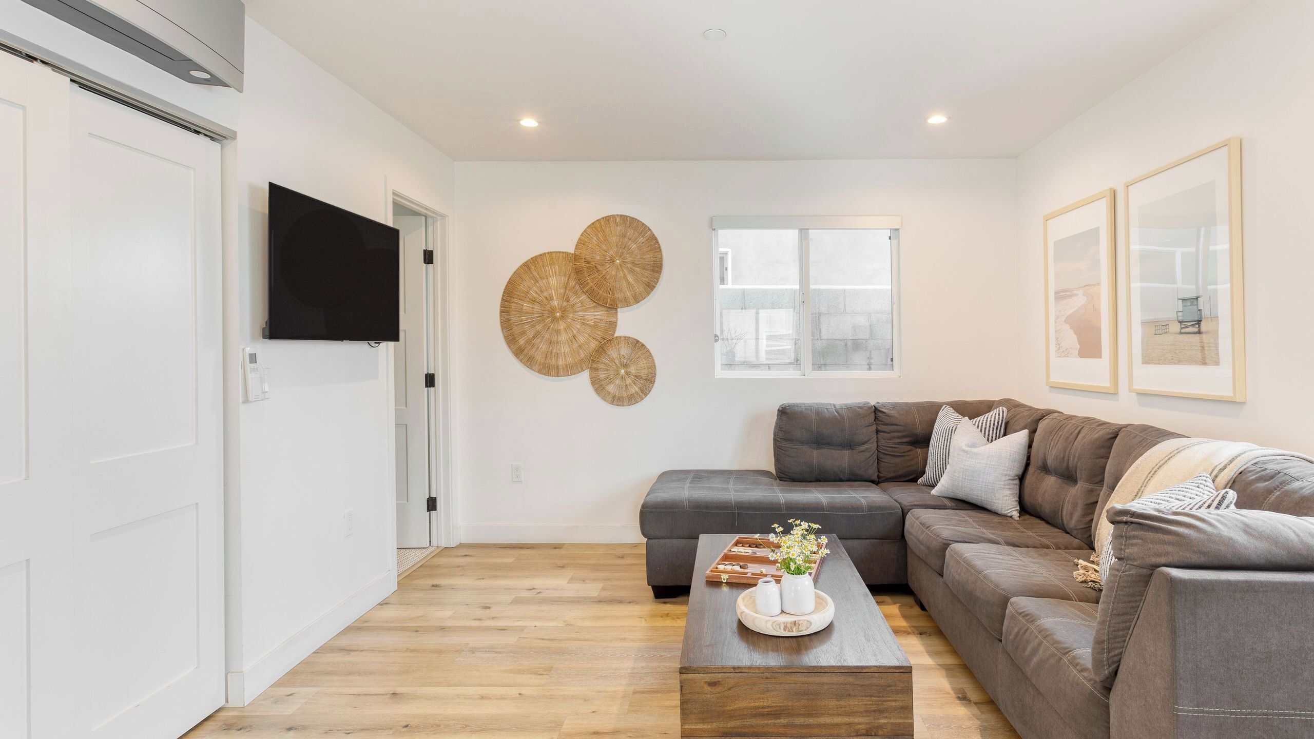 A modern living room with a sectional sofa, a coffee table with decorative items, a flat-screen TV mounted on the wall, and a window with a view.