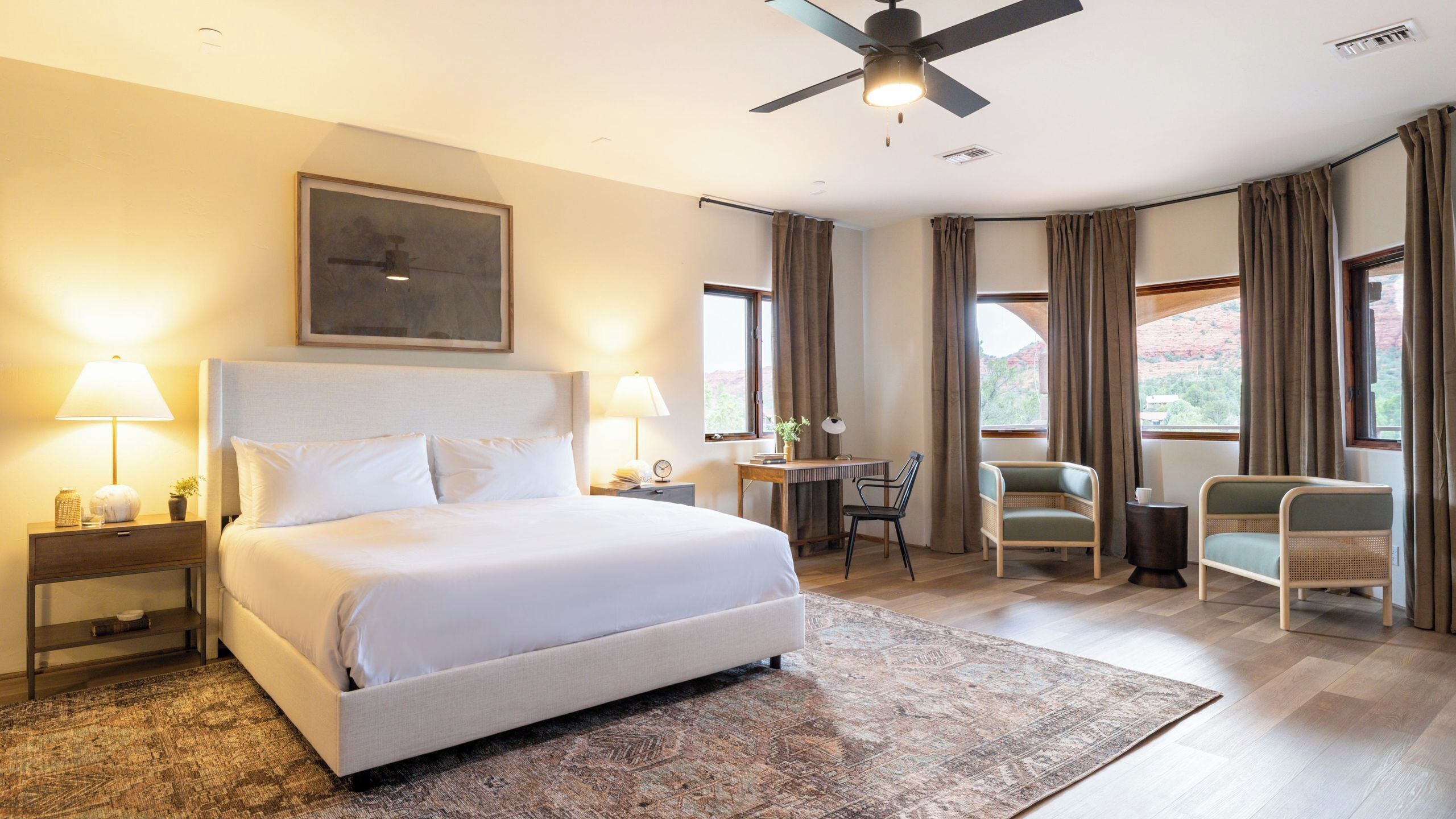 A hotel room with a bed, desk, two chairs, a ceiling fan, and large windows.