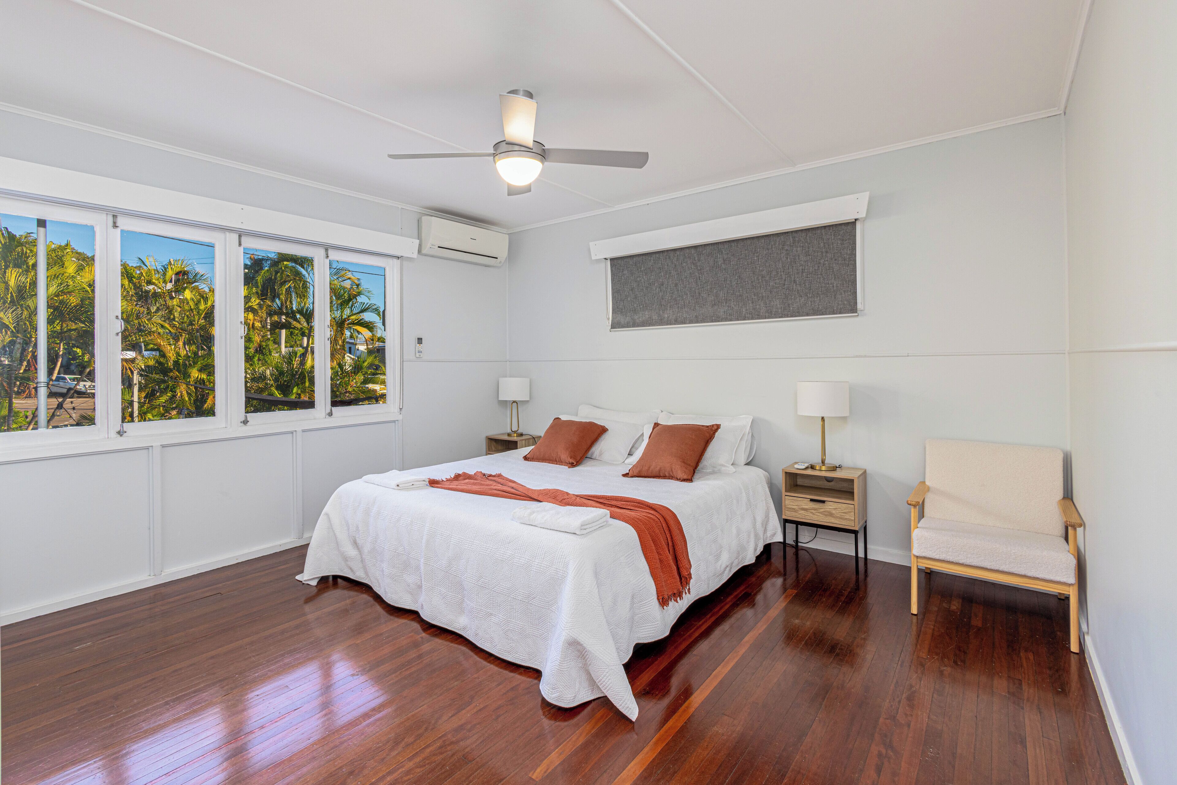 Une chambre avec un grand lit, un ventilateur de plafond, une fenêtre donnant sur des palmiers et un fauteuil.