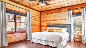 A wooden cabin bedroom with a bed, curtains, a ceiling fan, and a window.