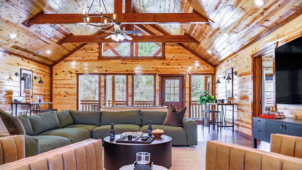 A log cabin interior with a large L-shaped sectional sofa, a round coffee table, and a television on the right.