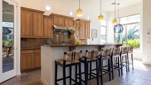 A kitchen with wooden cabinets, a central island with bar stools, pendant lighting, and a view of greenery outside.