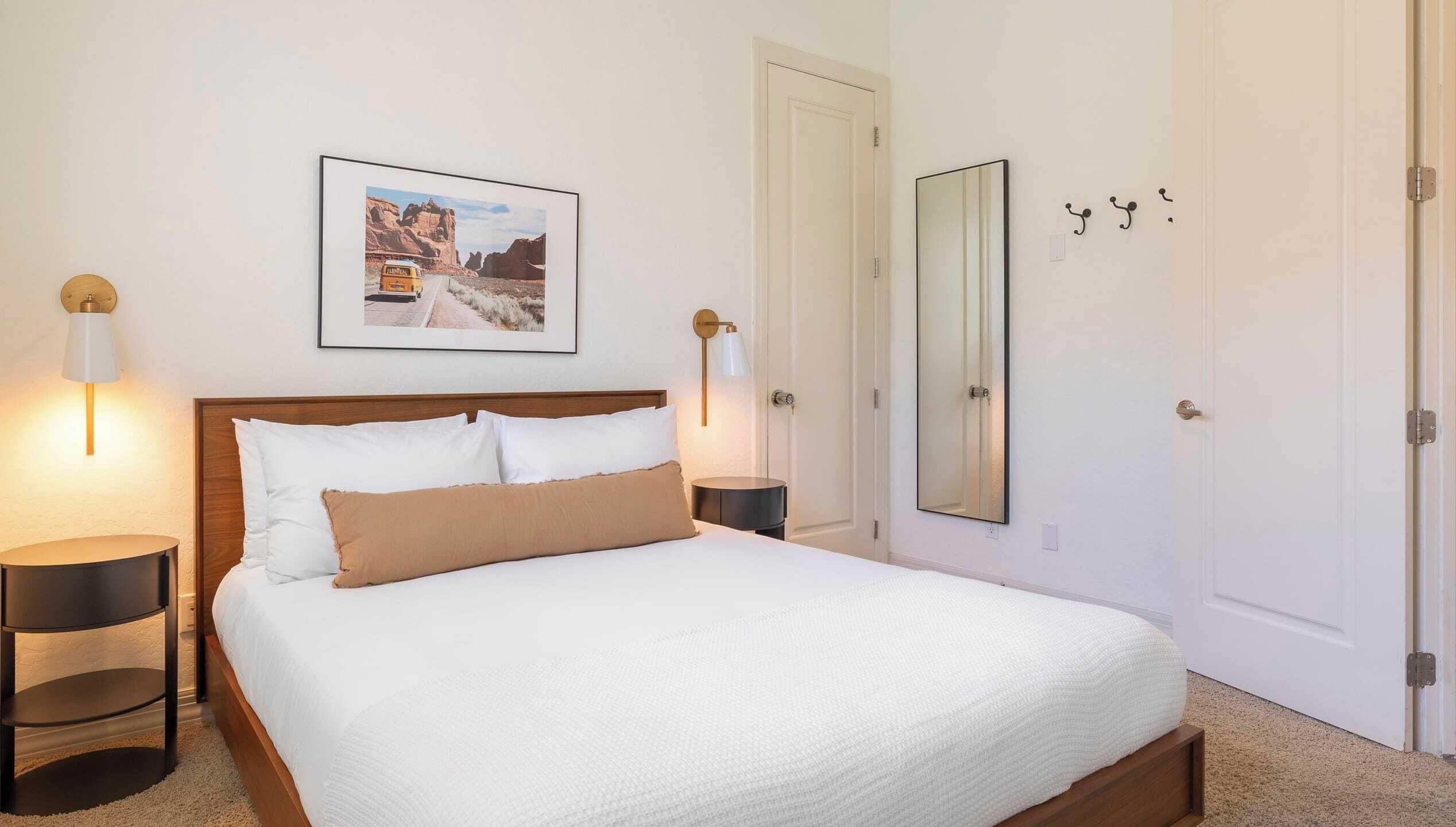 Une chambre à coucher bien rangée, avec un grand lit, deux tables de chevet, une lampe fixée au mur, un tableau encadré et une porte dotée d’un miroir.