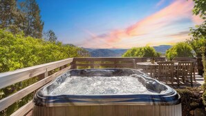 A hot tub on a wooden deck with a table and chairs, surrounded by greenery and a scenic view of mountains. - Asher by Avantstay Mountain Escape + Pool/spa (Weaverville)