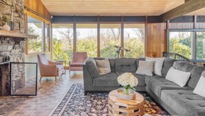 A cozy living room with a stone fireplace, large windows, a grey sectional sofa, and a small round coffee table. - Asher by Avantstay Mountain Escape + Pool/spa (Weaverville)