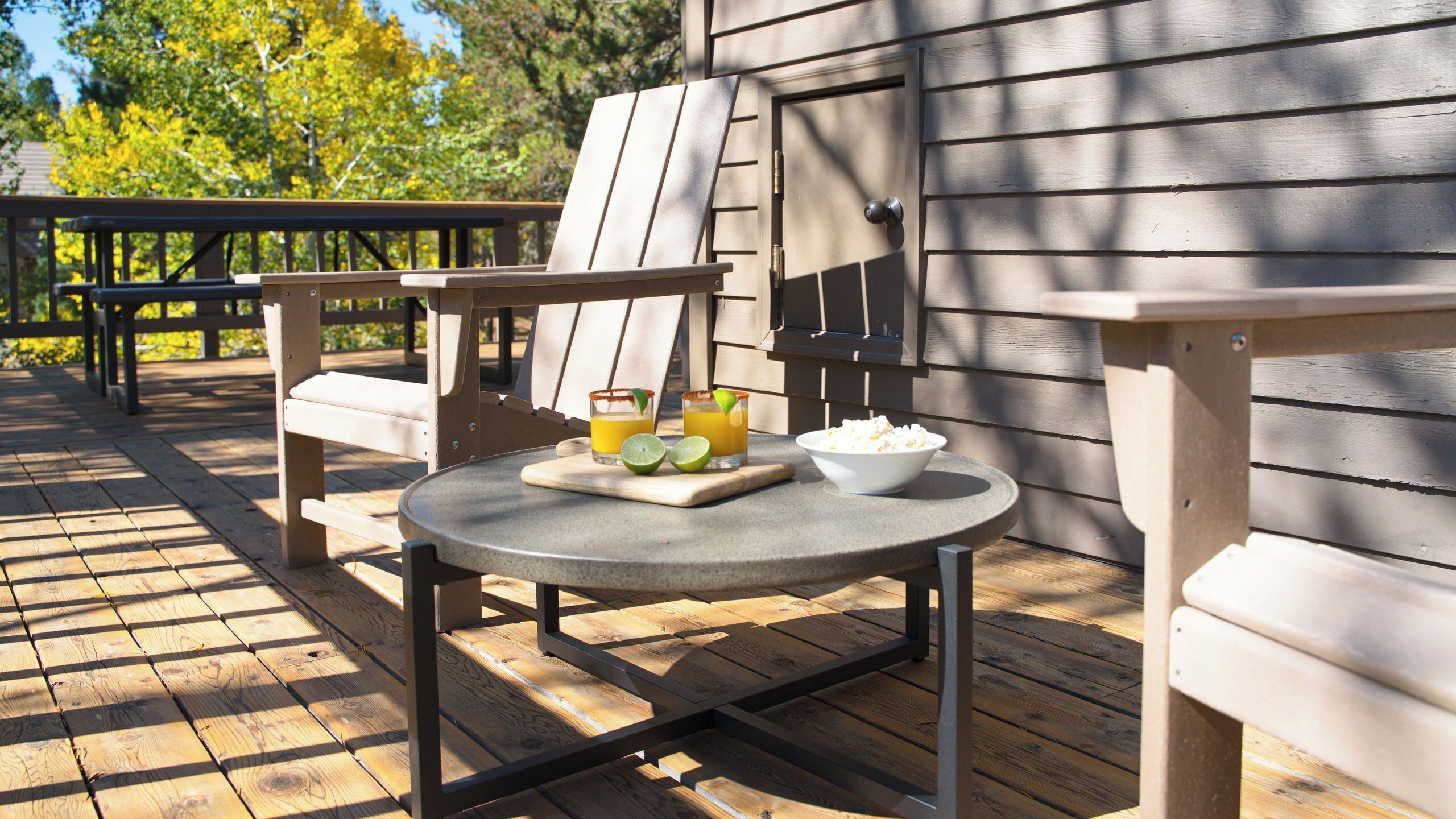 An outdoor patio with wooden furniture, a table with drinks and snacks, and a wooden building in the background.