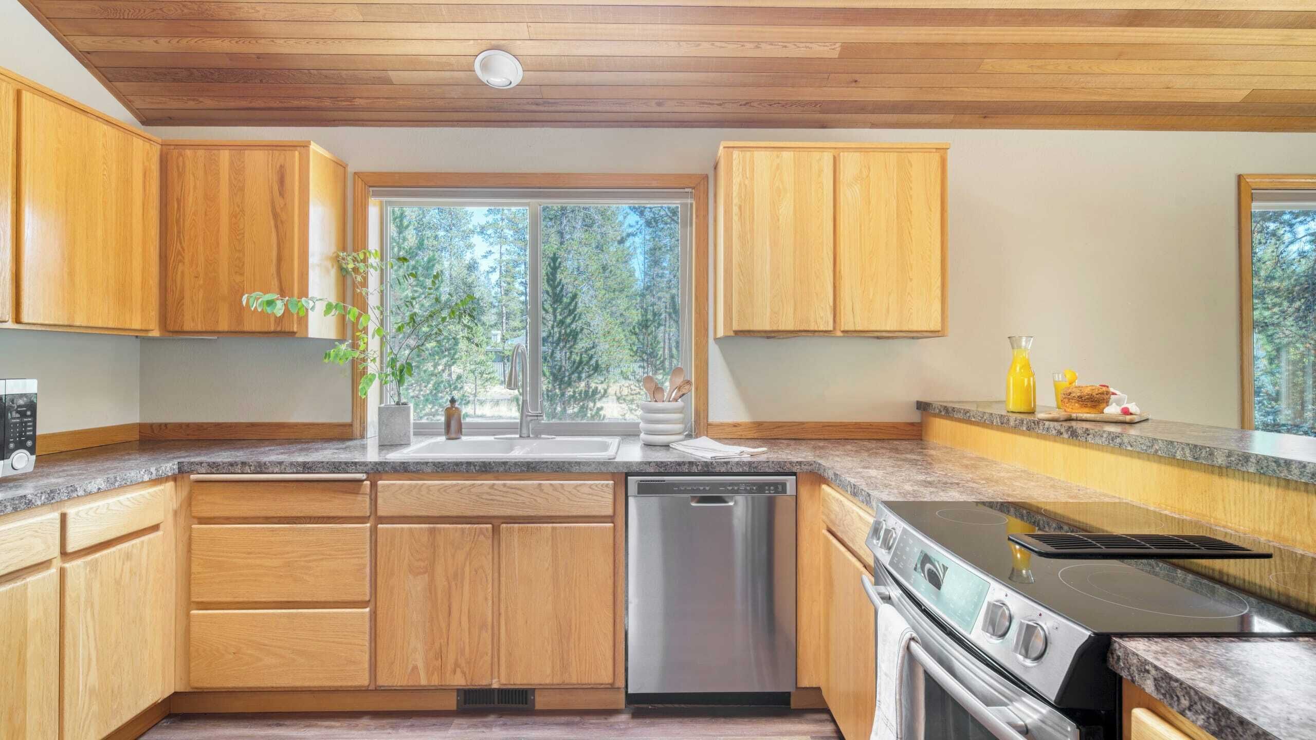 A modern kitchen with wooden cabinets, a stainless steel dishwasher, a built-in oven, and a large window offering a view of trees.