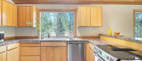 A modern kitchen with wooden cabinets, a stainless steel dishwasher, a built-in oven, and a large window offering a view of trees.