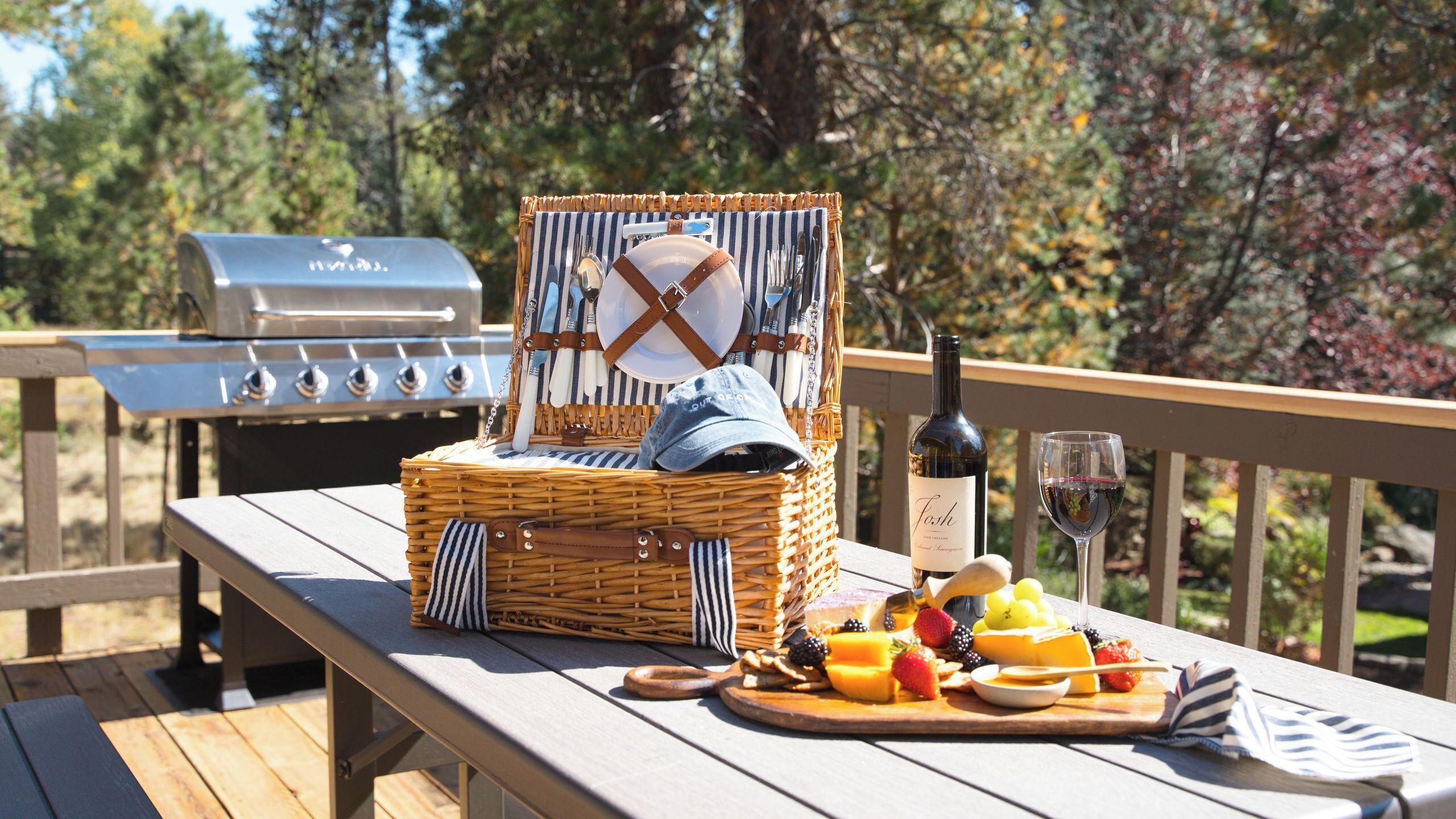 Preparación para un picnic con una cesta de mimbre, una botella de vino y una bandeja de frutas sobre una terraza de madera.